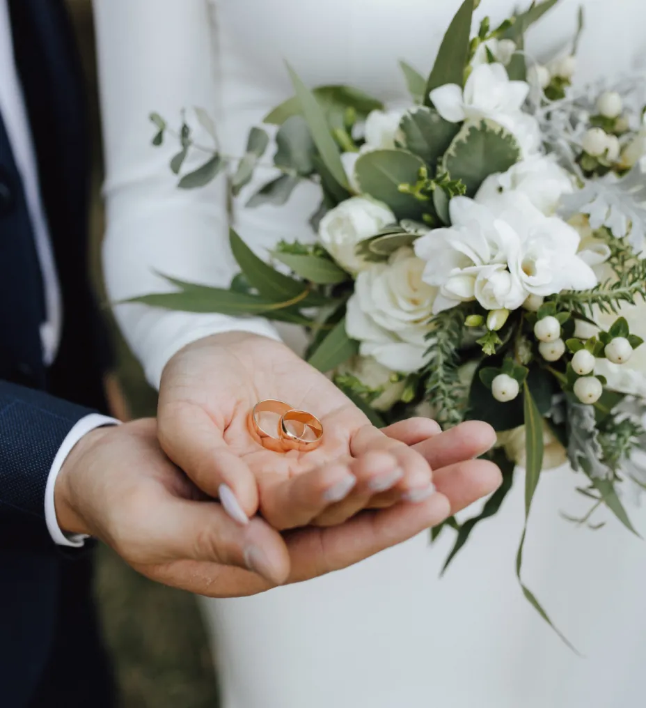 Un primo piano delle mani di una coppia che regge due fedi nuziali d'oro, accompagnate da un bellissimo bouquet di fiori bianchi e verde.